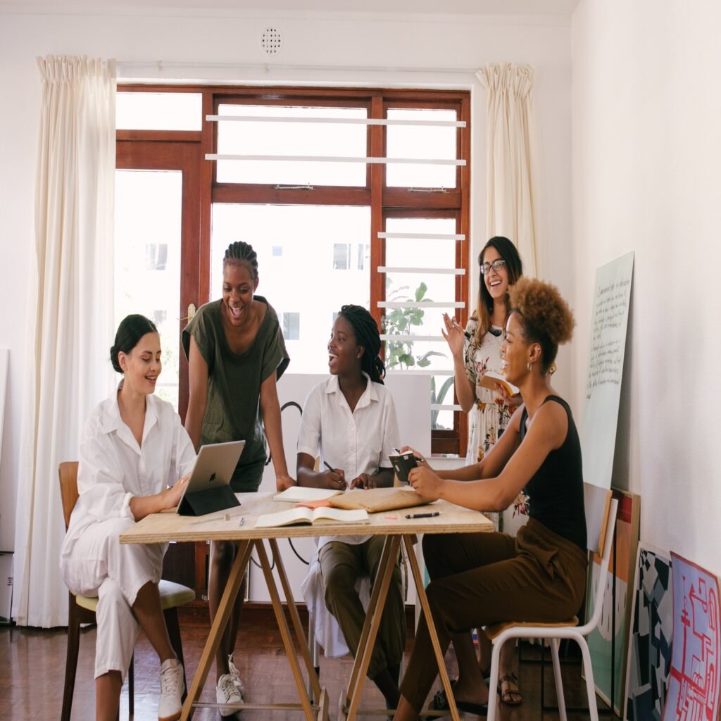 5 mulheres em um ambiente de trabalho, em volta de uma mesa, com livros, notebooks e uma lousa, como se estivessem elaborando estratégias