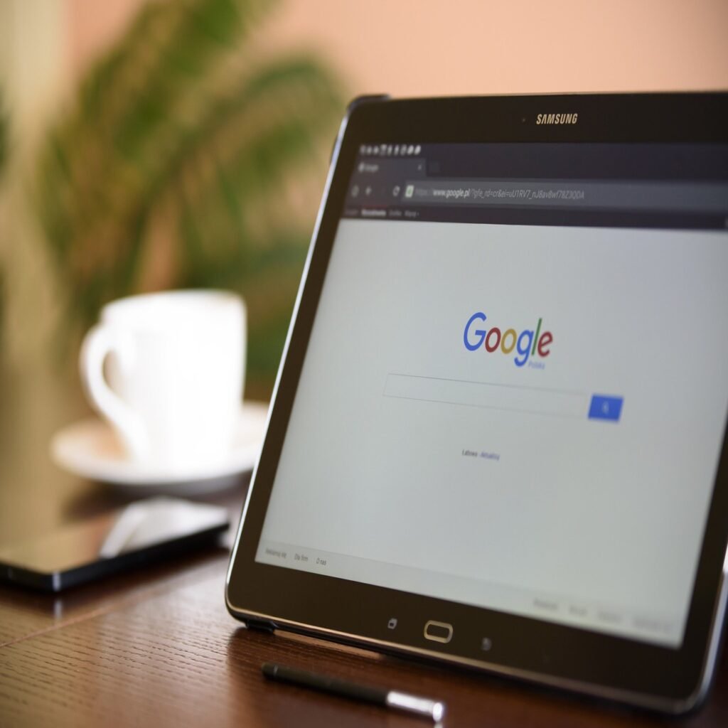 Tablet com a página inicial do Google Chrome aberta em cima de uma mesa de madeira. Ao fundo, pode-se ver um celular e uma xícara desfocados.