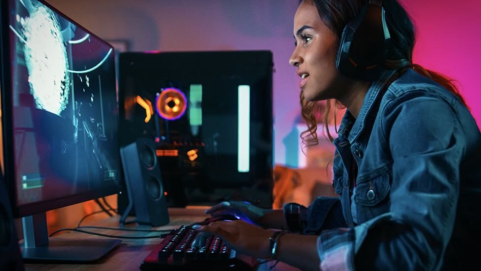 Na fotografia há uma garota negra de cabelos castanhos vestindo uma jaqueta jeans azul. Ela está em frente a um computador, por onde está jogando algum jogo eletrônico.