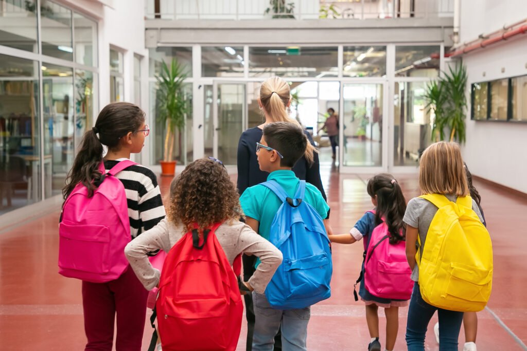 A imagem mostra cinco crianças, quatro meninas e um menino andando em direção à entrada da escola, guiados por uma mulher branca adulta.