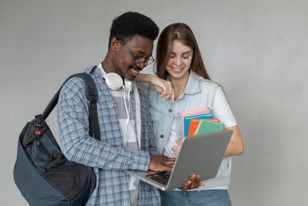 A imagem mostra dois jovens, um homem negro e uma mulher branca, parados em pé, segurando materiais de estudo (livros e mochila) e conversando enquanto olham e apontam para um computador.