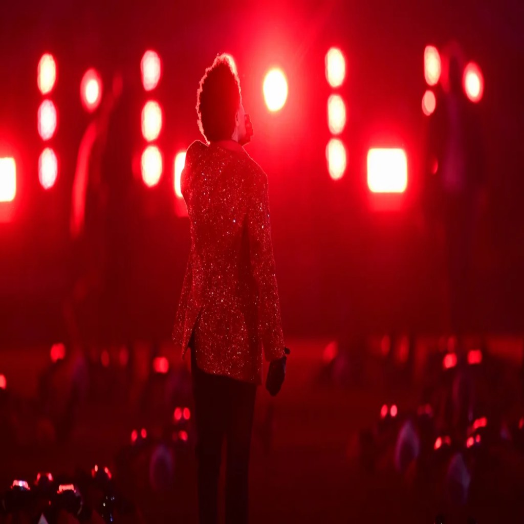 A foto mostra o cantor The Weeknd centralizado, com um black power, de costas para a foto. Ele está usando um terno vermelho brilhante, calças pretas e luvas pretas. Ao fundo, há inúmeros holofotes emitindo a luz vermelha, com a temática de seu álbum "Blinding Lights"