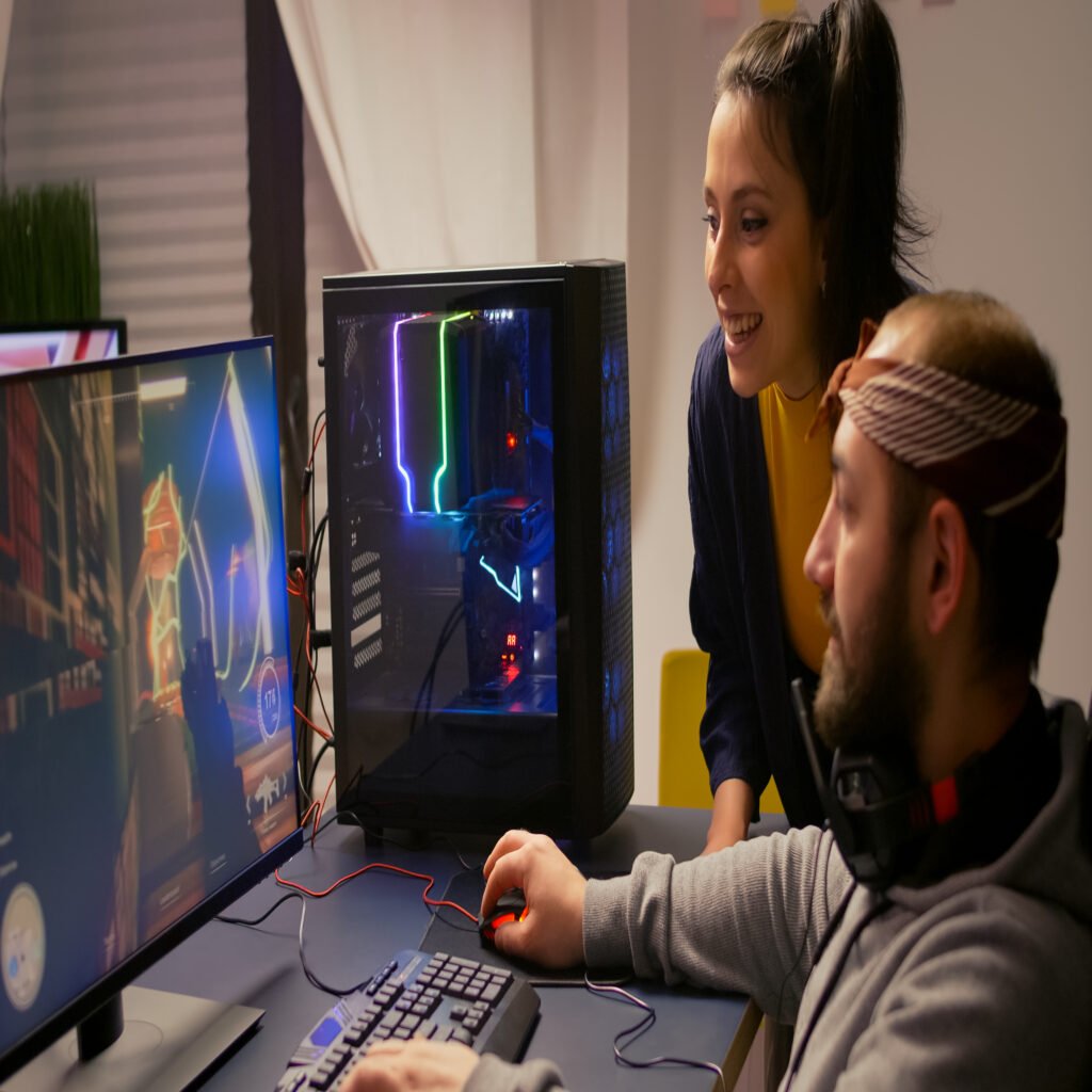 A imagem possui duas pessoas. Um homem branco, de barba e lenço na cabeça, e uma mulher branca de cabelo preso em um rabo de cavalo. Os dois sorriem enquanto olham para tela do computador onde estão jogando um jogo.