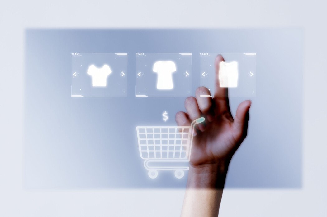 A imagem possui um fundo azul claro, com uma mão humana clicando ícones de peças de roupas de maneira virtual, em uma base transparente. Abaixo, possui um carrinho de compras.