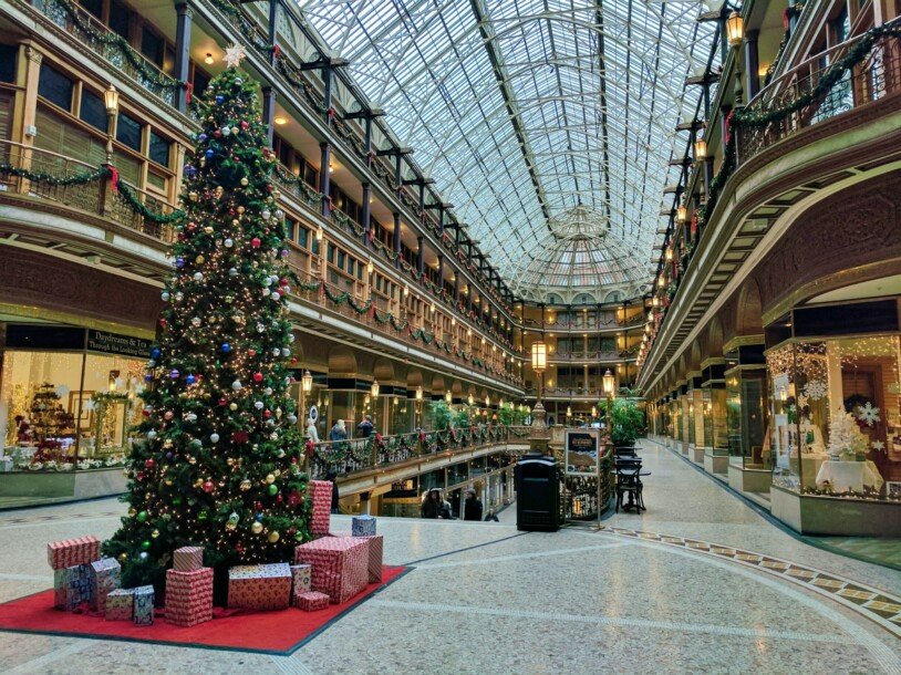 Um shopping, com as lojas com uma decoração natalina, flocos de neve e faixas de decoração verde no apoio dos corredores. Na parte esquerda da imagem há uma árvore de natal decorada, com uma estrela no topo e embalagens de presente ao seu redor no chão